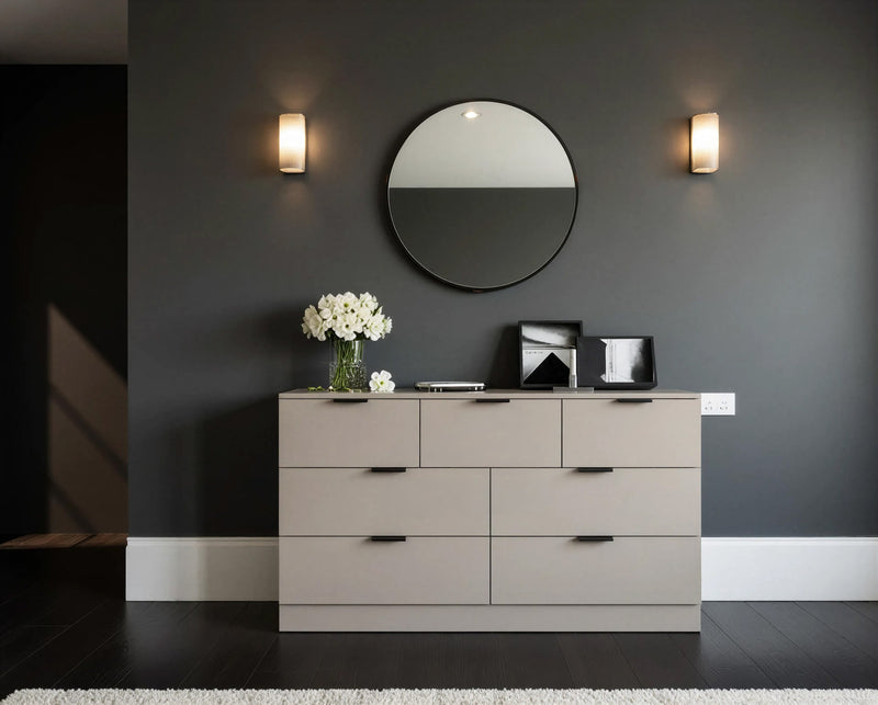 Battersea chest of drawers: a wide seven-drawer chest in warm greige with black bar handles under a round mirror on a dark wall