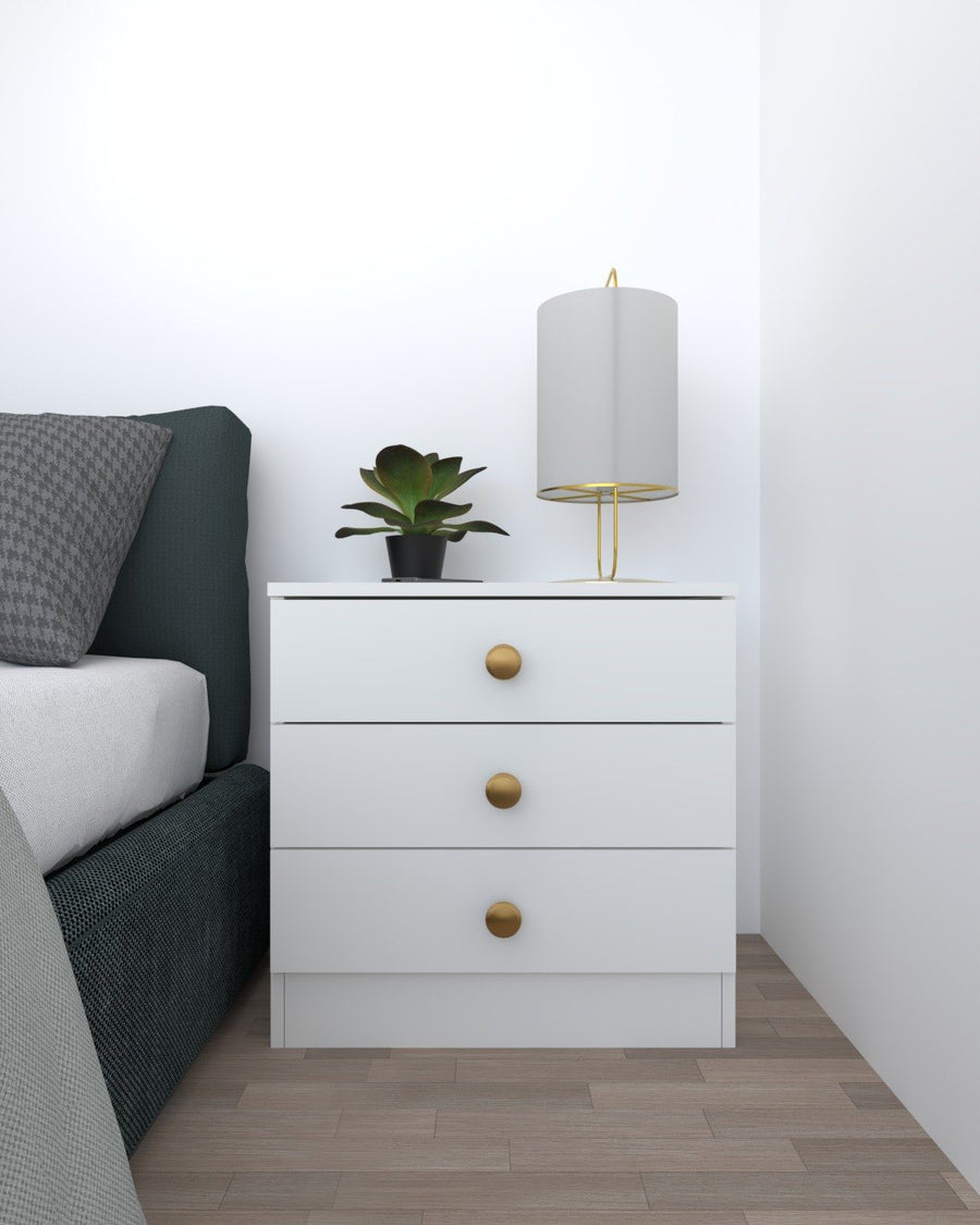White three-drawer bedside table with brass knobs beside a bed, with a lamp and a plant on top