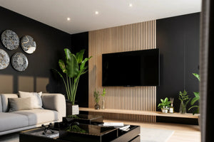 Hertford oak slatted TV wall panel in London: a floor-to-ceiling pale oak slat panel set into a charcoal wall, with the television on it and a long floating oak shelf beneath, seen from the sofa. Made to measure by Metro Home Interiors