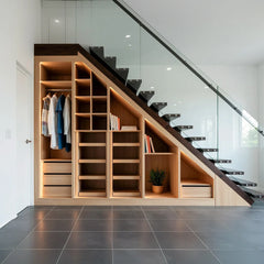 Hoxton under-stairs wardrobe: an open oak cloakroom beneath a floating staircase with a hanging rail, pigeonhole shelves, a drawer bank and LED lighting on a dark tiled floor