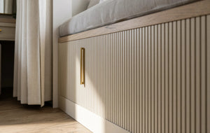 Seraphina fitted bedroom furniture in London: Cashmere Grey shaker wardrobes with brass bar handles; the fluted panel and brass lighting strip under the window seat. Made to measure by Metro Home Interiors